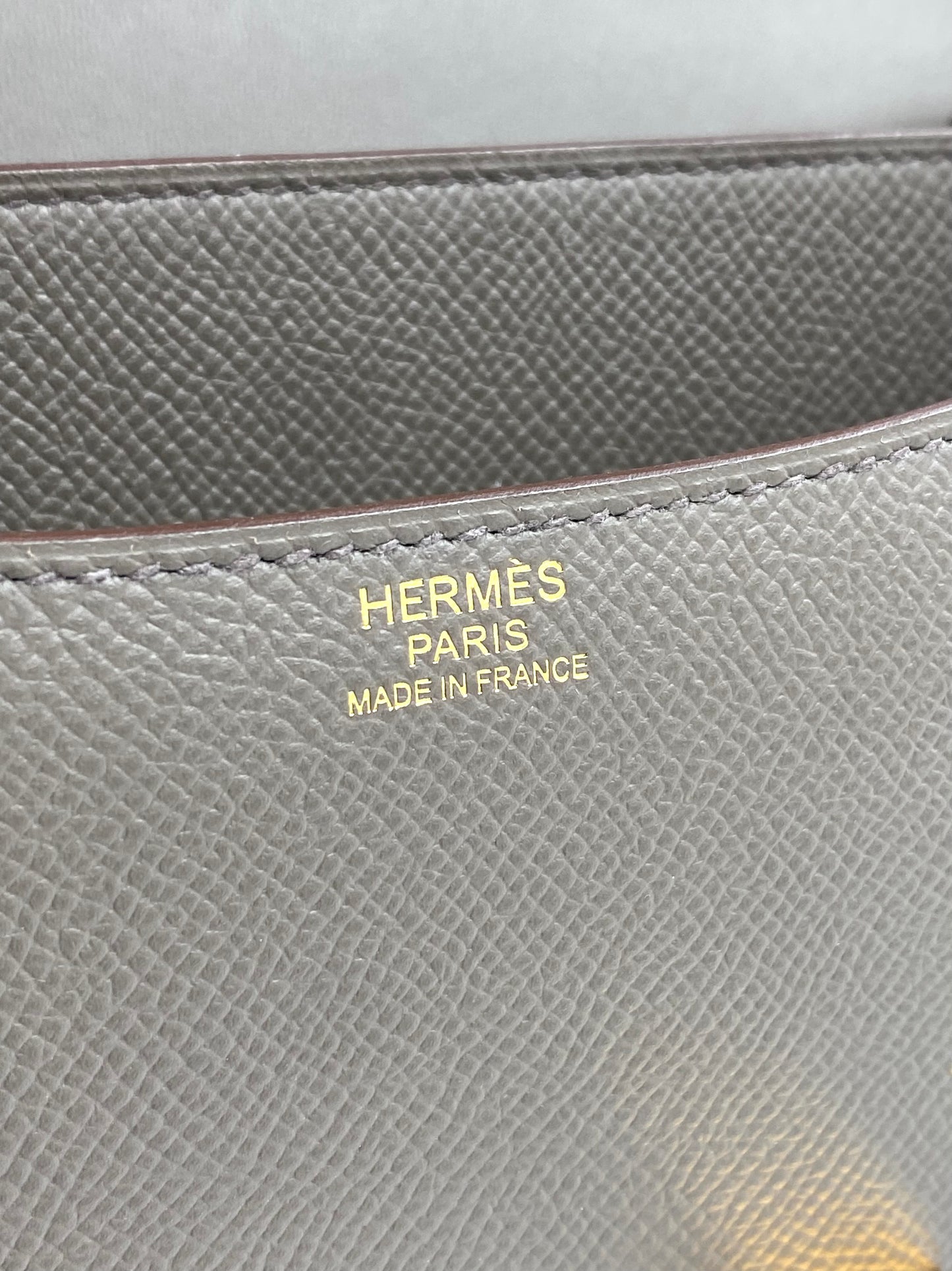 HERMÈS Constance 24 Etain Epsom Roségold Hardware Fullset mit Original Rechnung Nahaufnahme des goldenen Prägeaufschritts auf grauem Epsomleder.