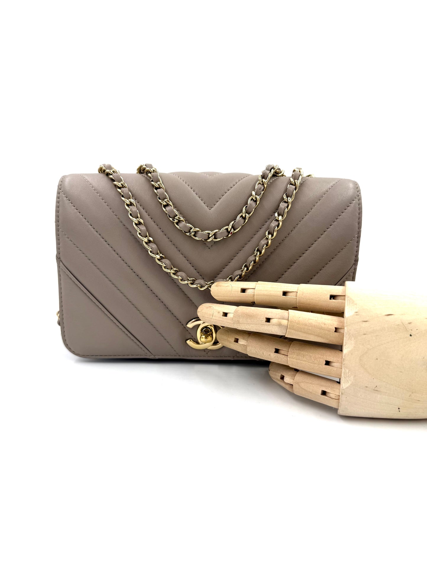 CHANEL Timeless Single Flap Bag Chevron taupe GHW mit goldfarbenem Kettenriemen auf weißem Hintergrund und Holz-Handmodell