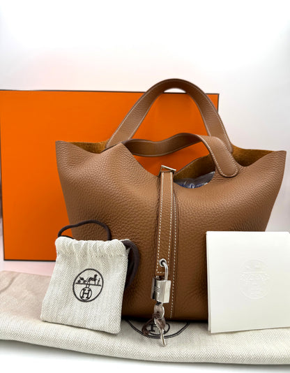 HERMÈS Picotin 26 Lock Clemence gold PHW Handtasche mit Schlüssel und Staubbeutel vor orangefarbenem Hintergrund