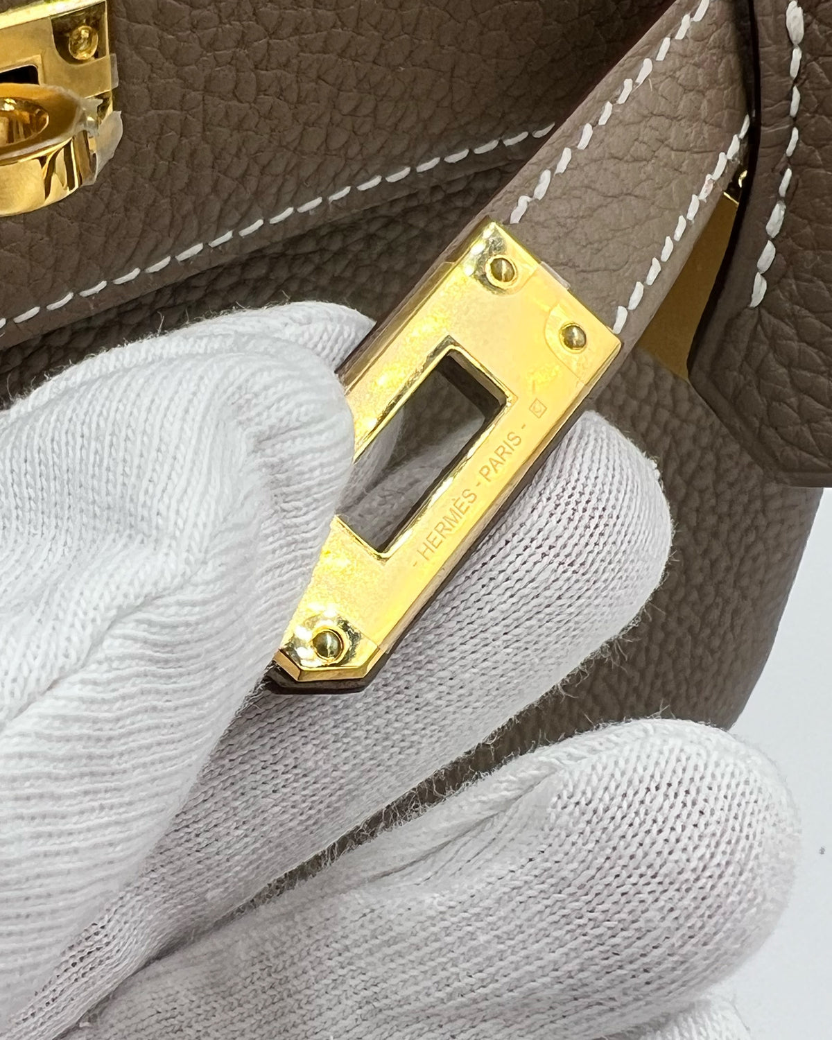 Detailaufnahme der HERMÈS Kelly 25 Togo Etoupe GHW mit goldfarbenem Verschluss und weißen Handschuhen