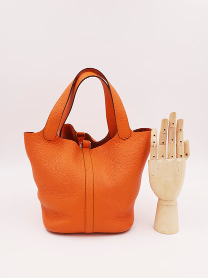 HERMÈS Picotin 22 orange PHW, orangefarbene Designerhandtasche aus Leder neben einer Holzhandskulptur