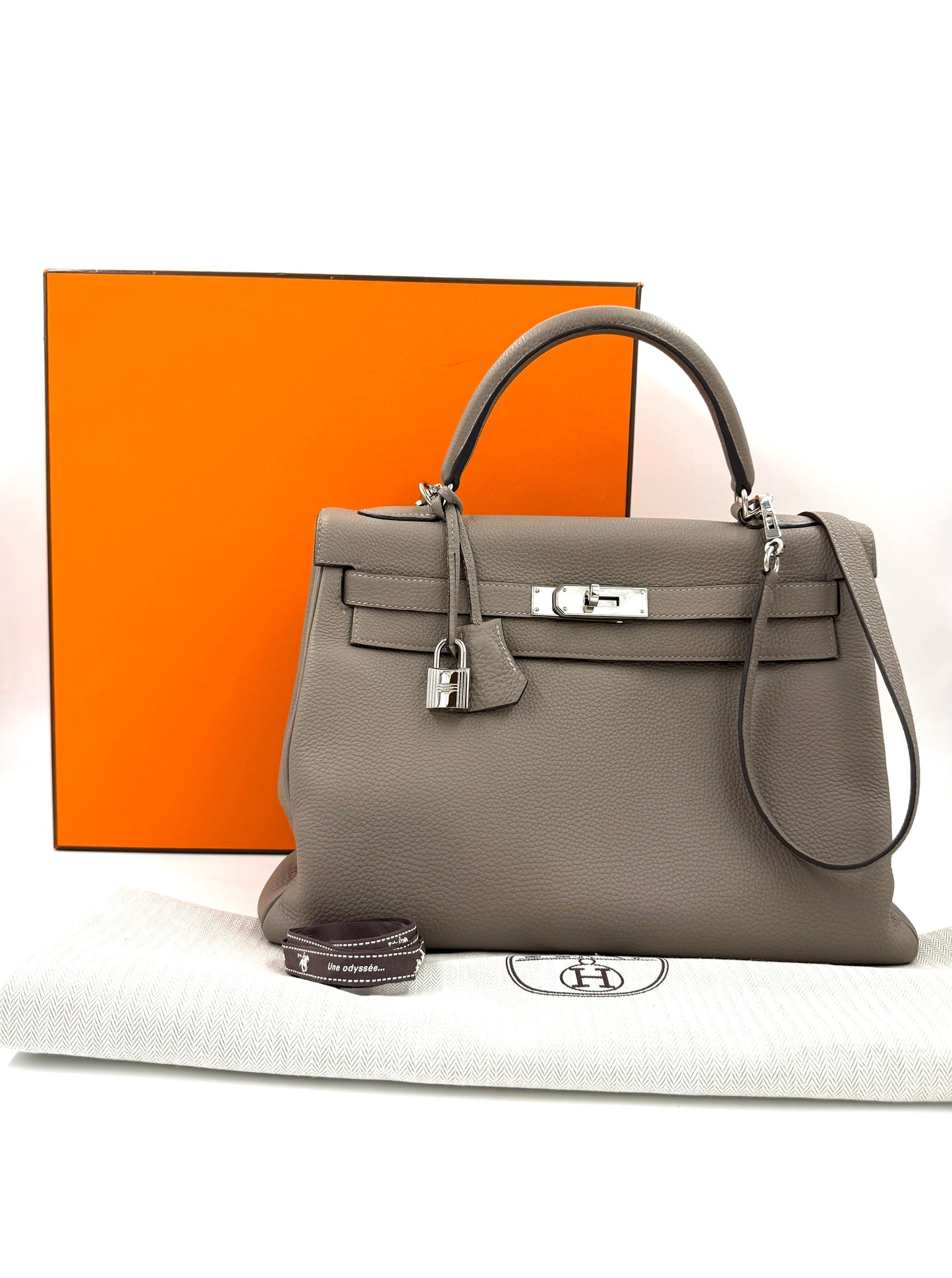 HERMÈS Kelly 32 Retourne Gris Asphalte PHW aus kalbsleder mit Palladium-Hardware und Staubbeutel vor orangenem Hermes-Karton.