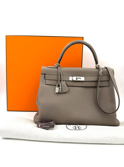 HERMÈS Kelly 32 Retourne Gris Asphalte PHW aus kalbsleder mit Palladium-Hardware und Staubbeutel vor orangenem Hermes-Karton.
