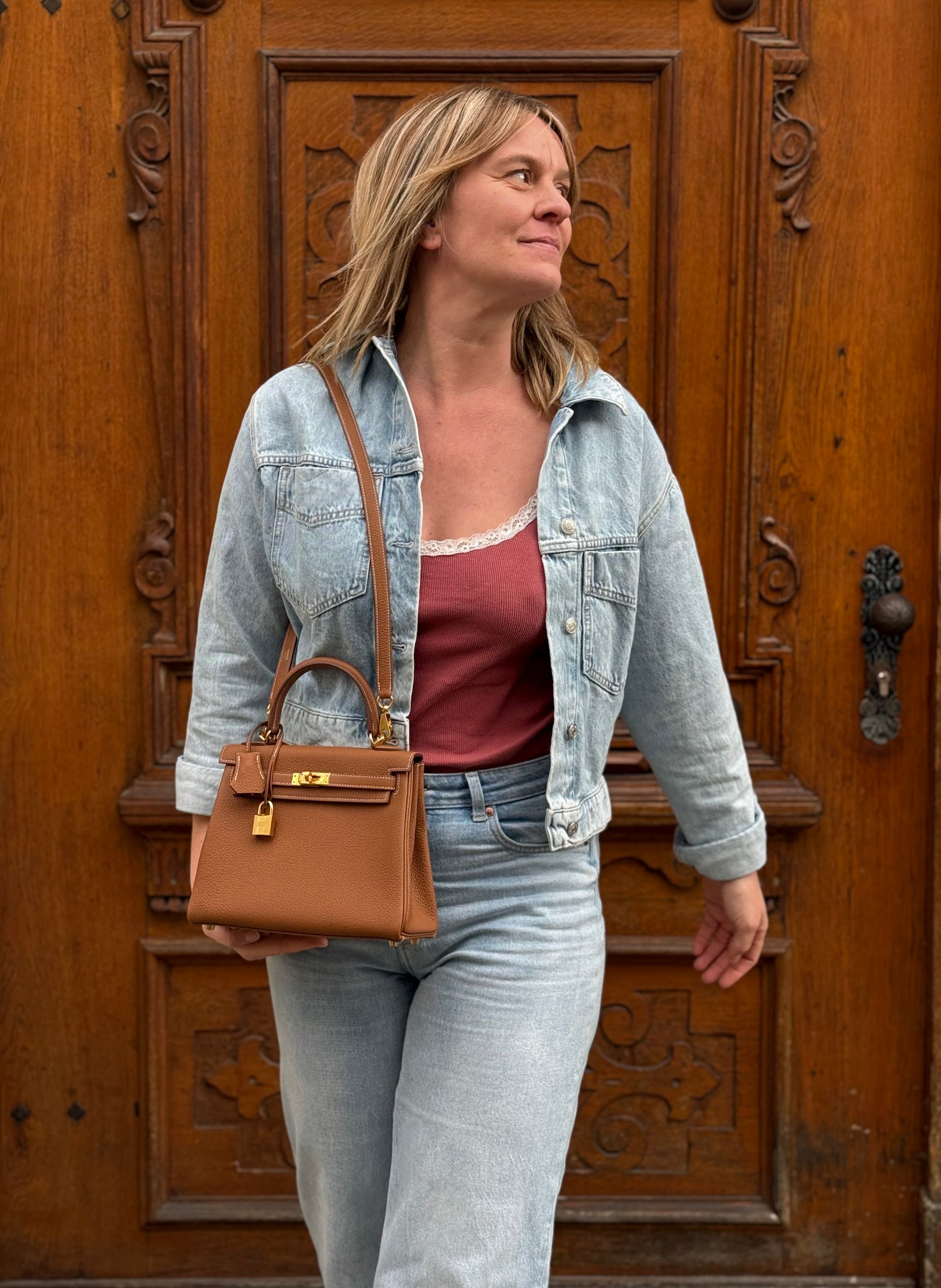 Frau mit blonden Haaren trägt hellblaue Jeansjacke, rotes Top und braune Lederhandtasche vor verzierter Holztür