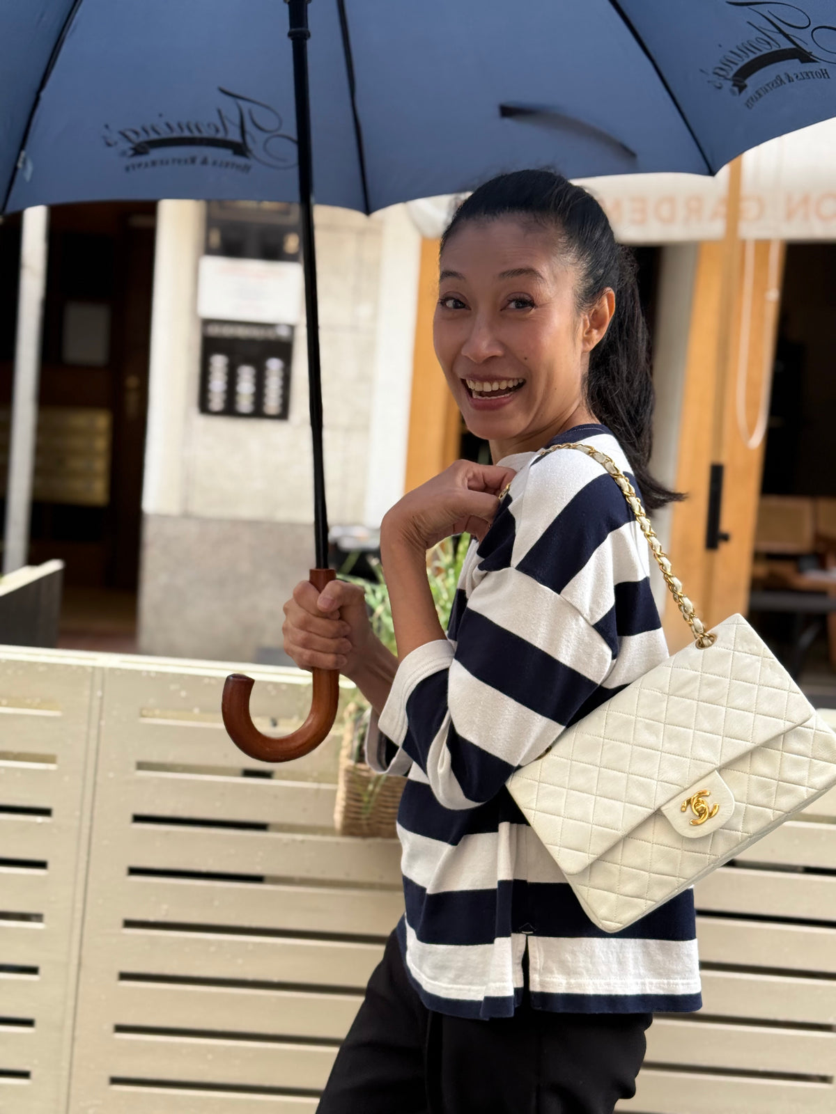 Frau mit gestreiftem Oberteil trägt eine weiße gesteppte Chanel-Handtasche unter einem Regenschirm.