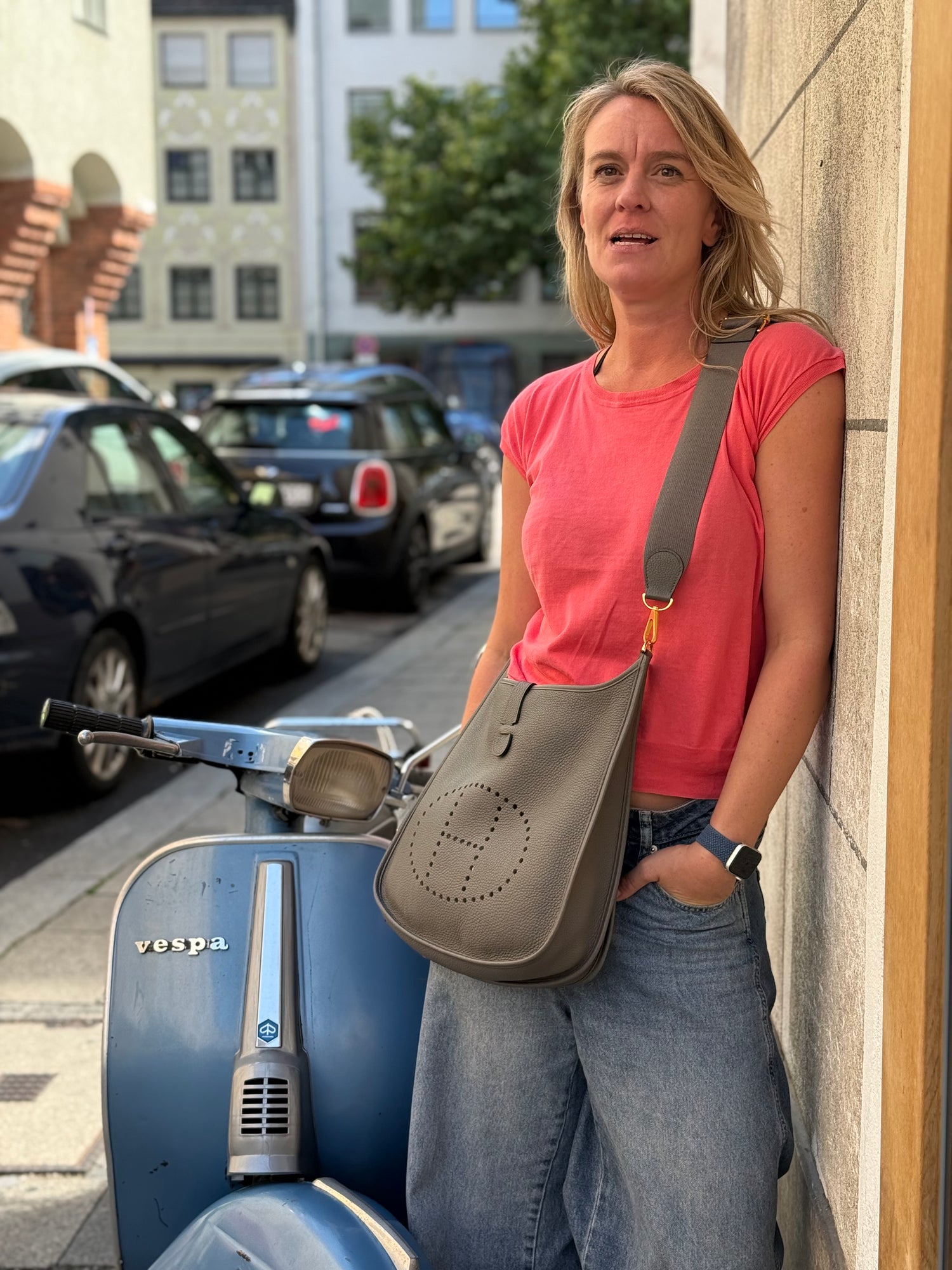 Frau mit grauer Umhängetasche neben einer blauen Vespa in einer belebten Straße