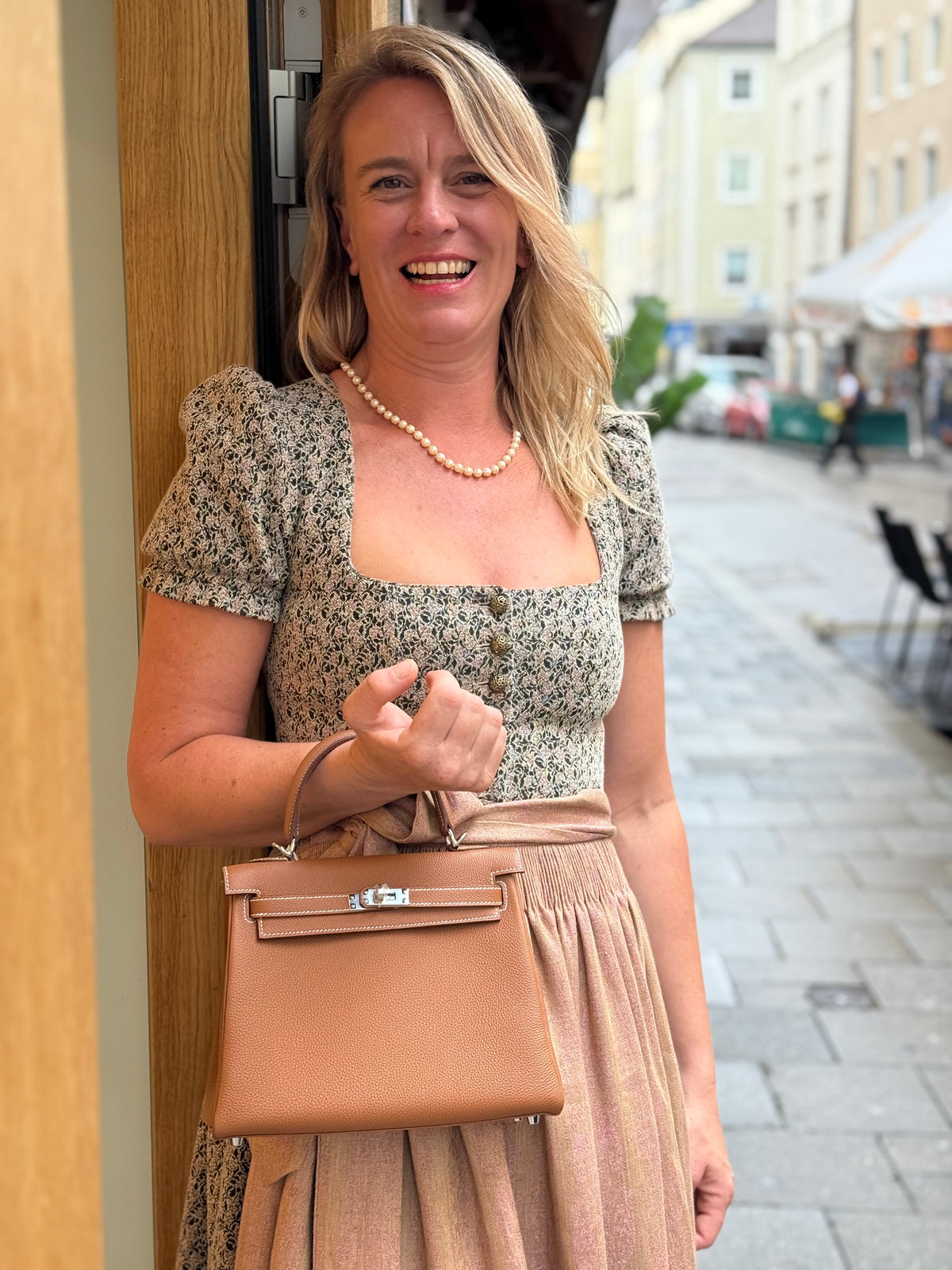 Lächelnde Frau mit eleganter brauner Handtasche vor einer Boutique in einer Münchner Altstadtgasse