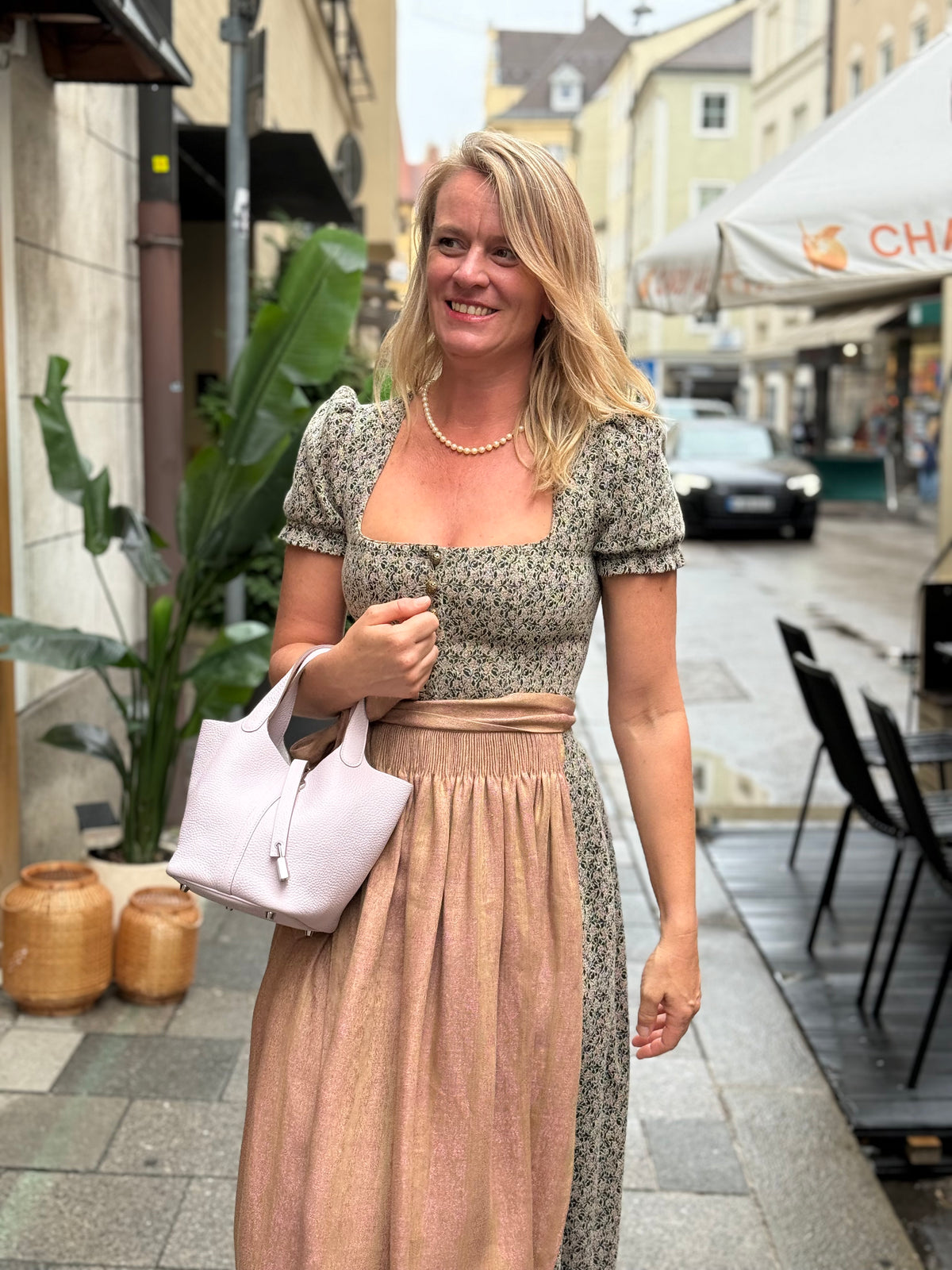 Lächelnde Frau im Dirndl mit heller Handtasche auf einer belebten Straße in München