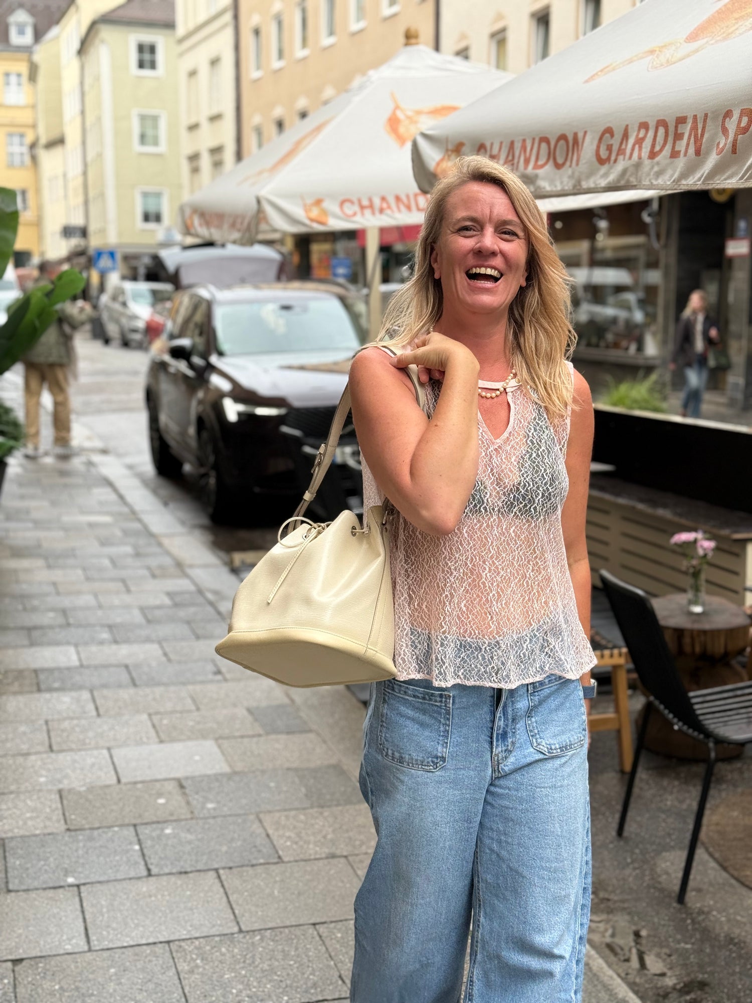 Lächelnde Frau mit heller Handtasche auf einer belebten Straße mit Cafés und Altstadthäusern