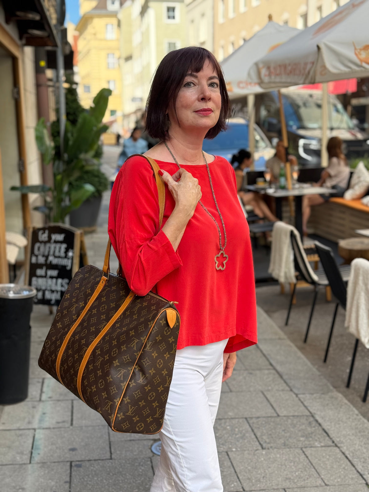 Frau mit Louis Vuitton Monogramm Tasche in Rot und Weiß vor einem Straßencafé in München