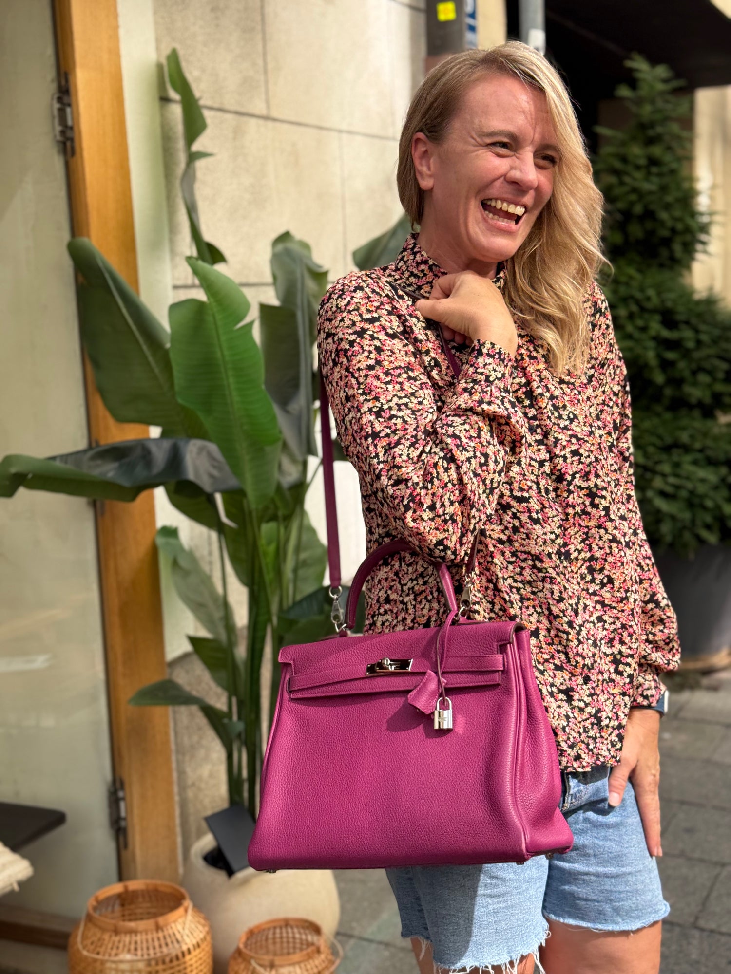 Lachende Frau mit magenta Handtasche, Blumenbluse und Jeansshorts vor einer Tür mit Pflanzen