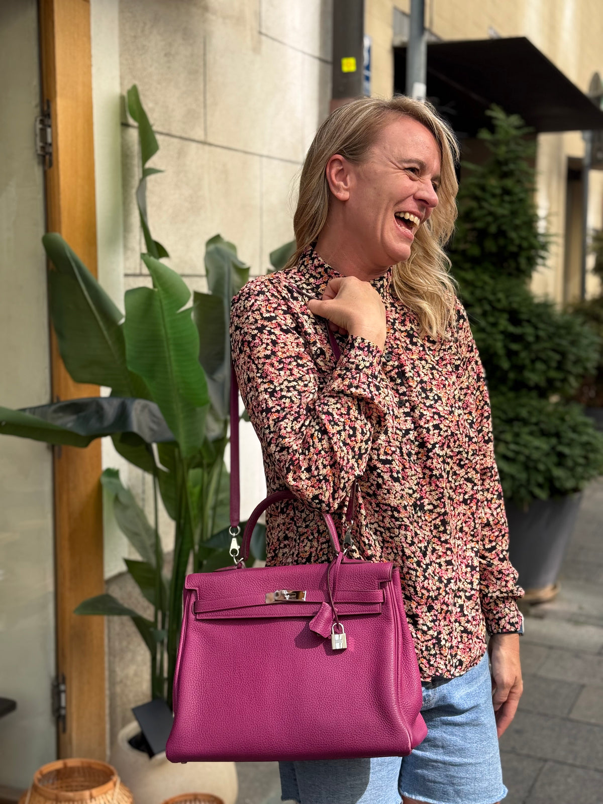 Lachende Frau mit pinker Handtasche vor einem Gebäude mit üppigen Pflanzen