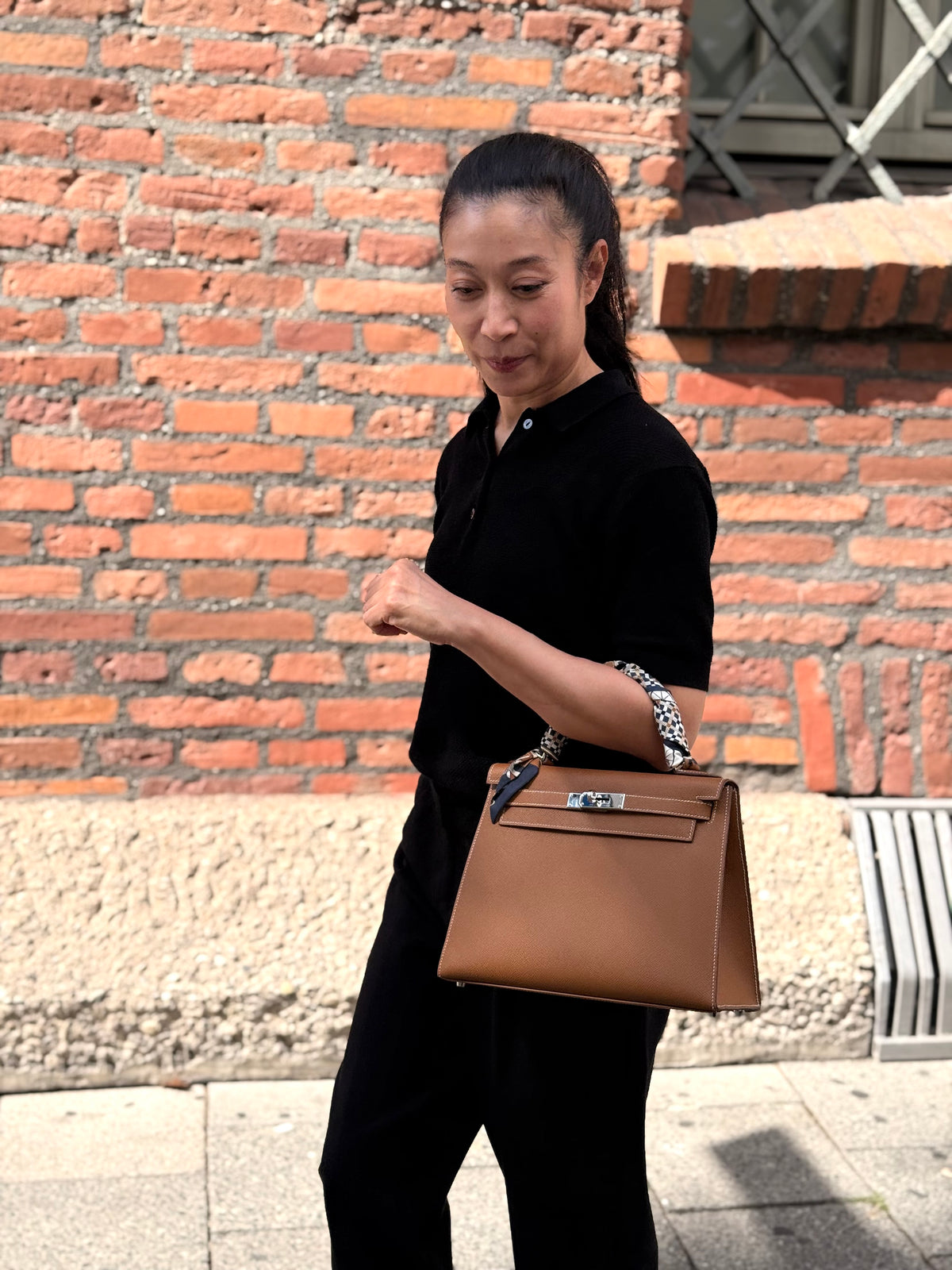 Frau in schwarzer Kleidung trägt eine braune Handtasche vor einer Backsteinwand.