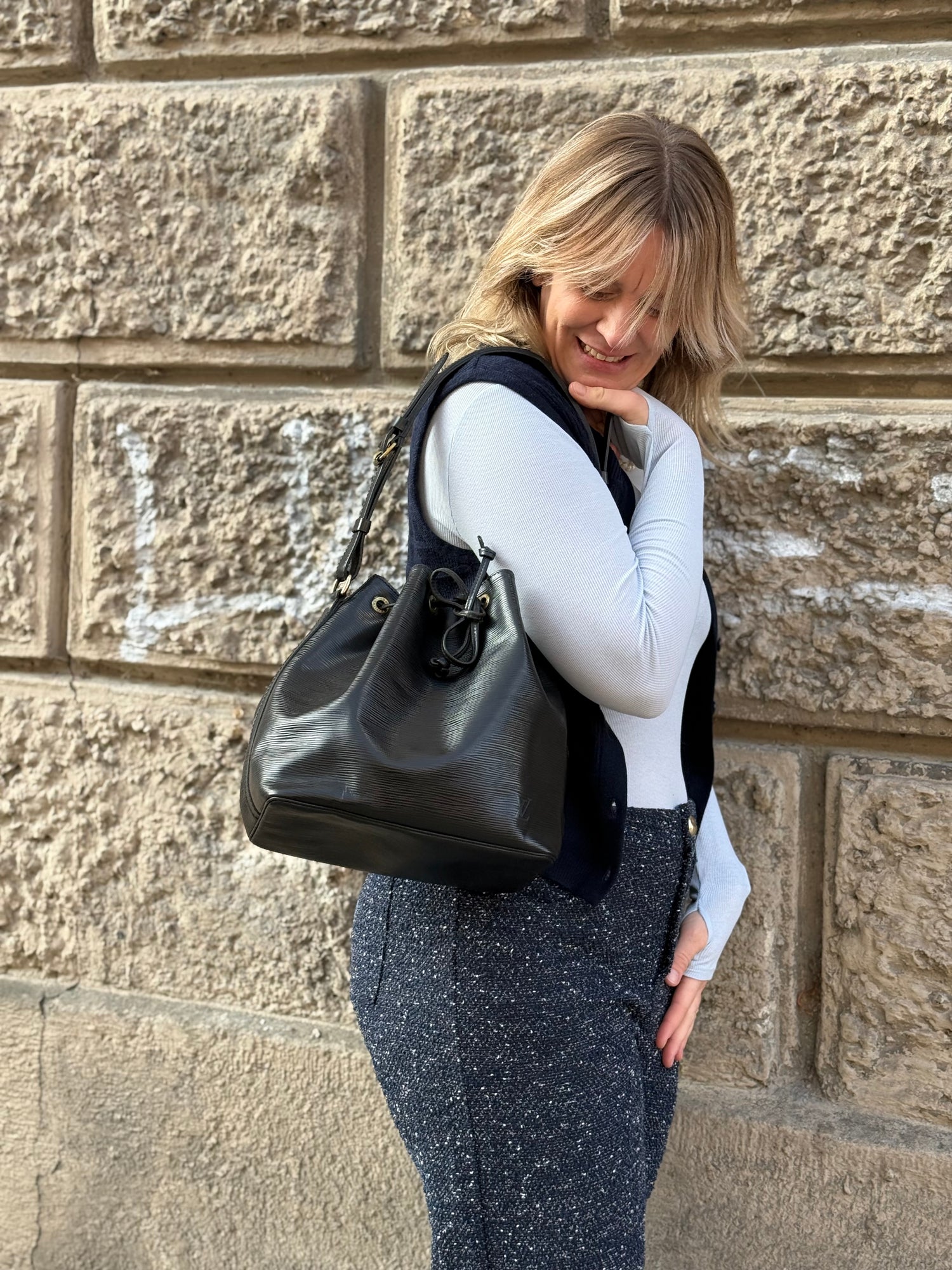 Frau mit hellblondem Haar trägt weißen Rollkragenpullover und schwarze Louis Vuitton Tasche vor Steinwand