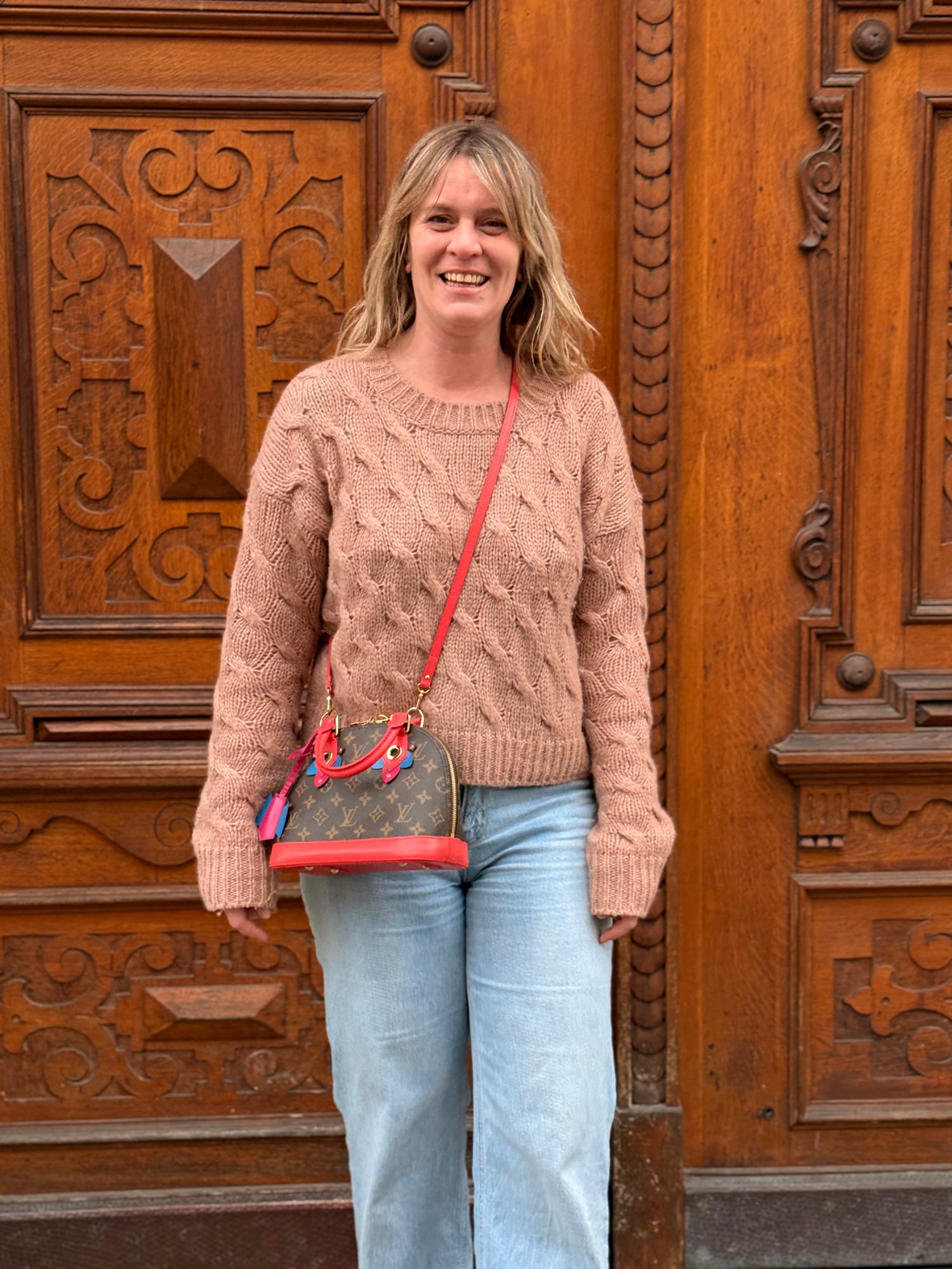 Frau mit beigen Strickpullover und Jeans trägt Louis Vuitton Tasche mit rotem Trageriemen vor holzgeschnitzter Tür