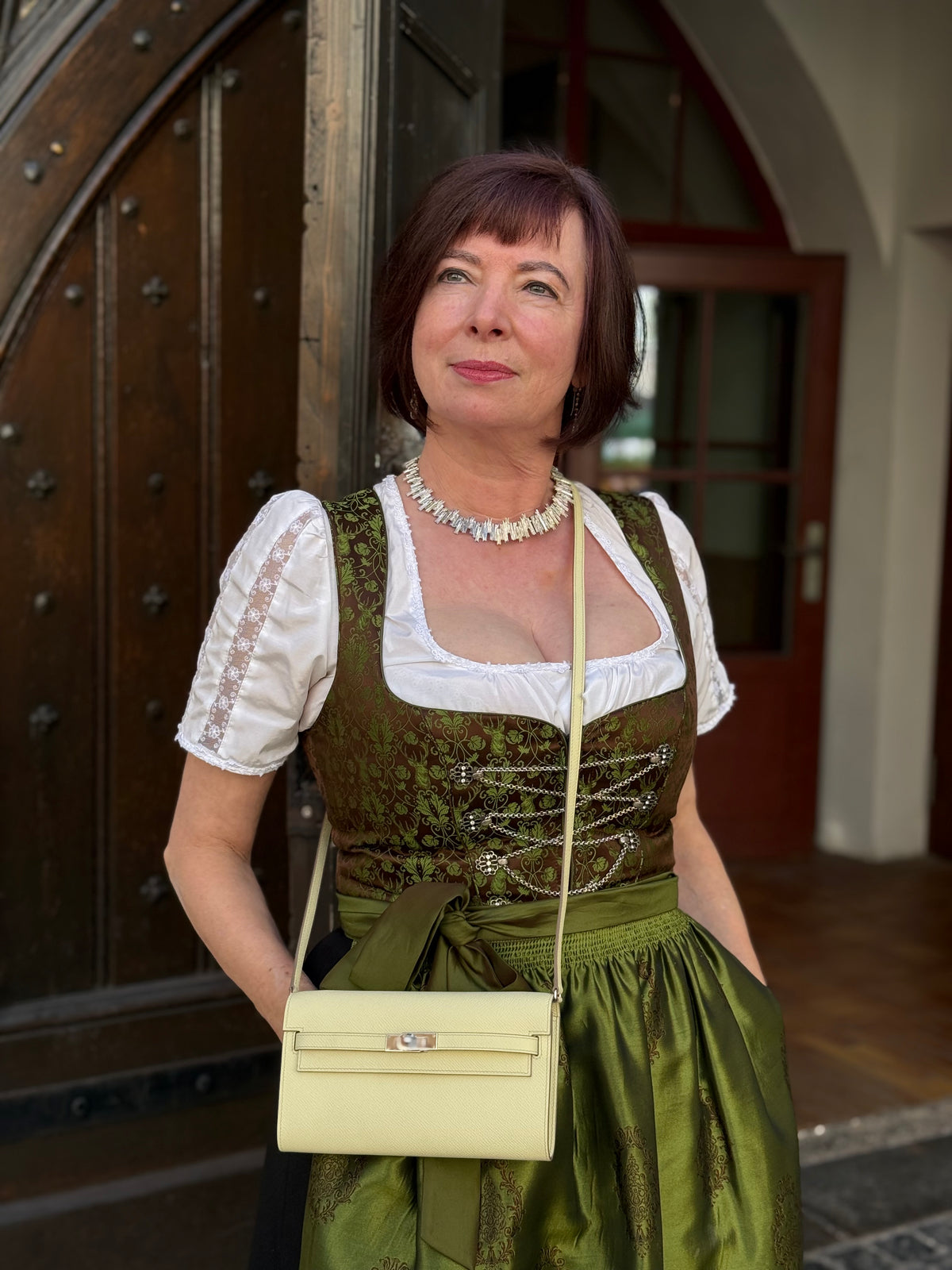 Frau im grünen Dirndl mit cremefarbener Handtasche vor einer historischen Holztür