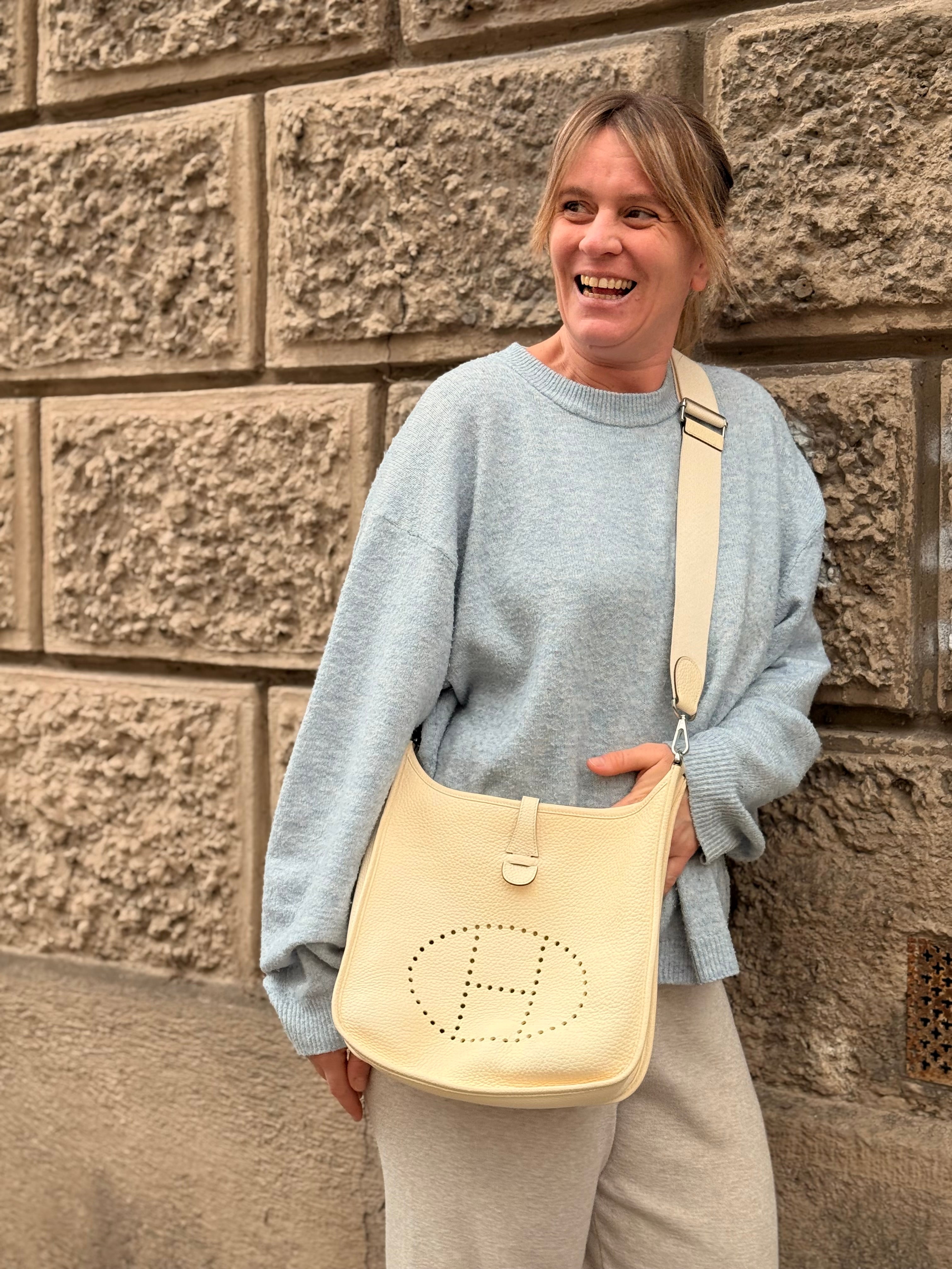 Frau mit hellgrauem Pullover trägt beige Hermès Umhängetasche mit markantem H-Logo vor raue Steinwand