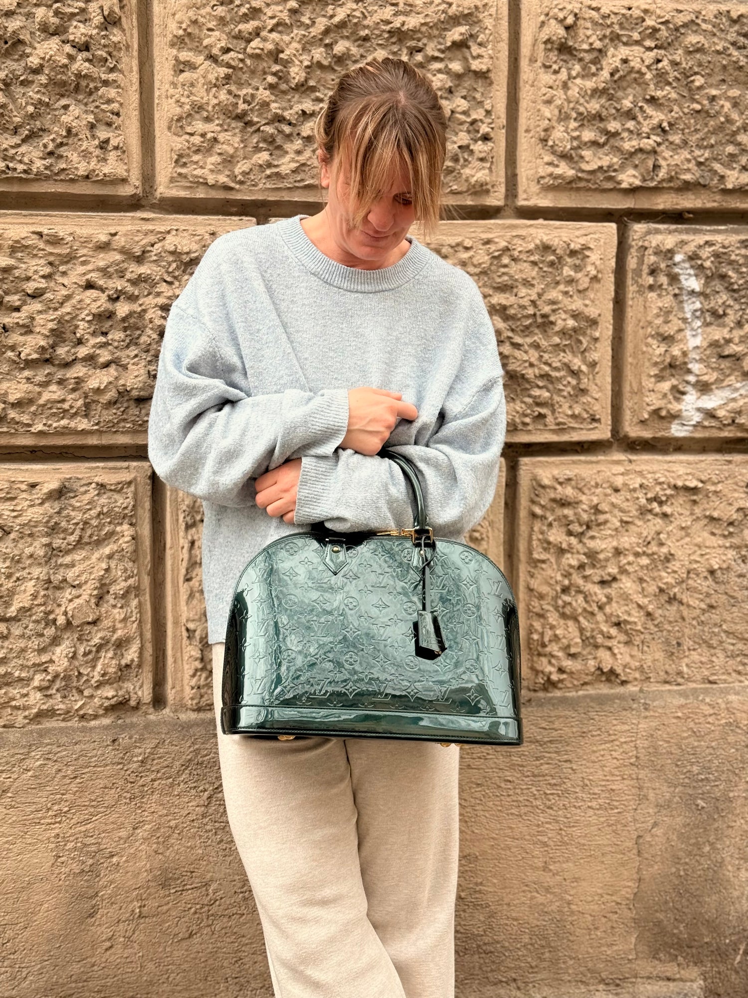 Frau in hellem Pullover und beiger Hose hält glänzende grüne Designertasche vor strukturierter Sandsteinwand