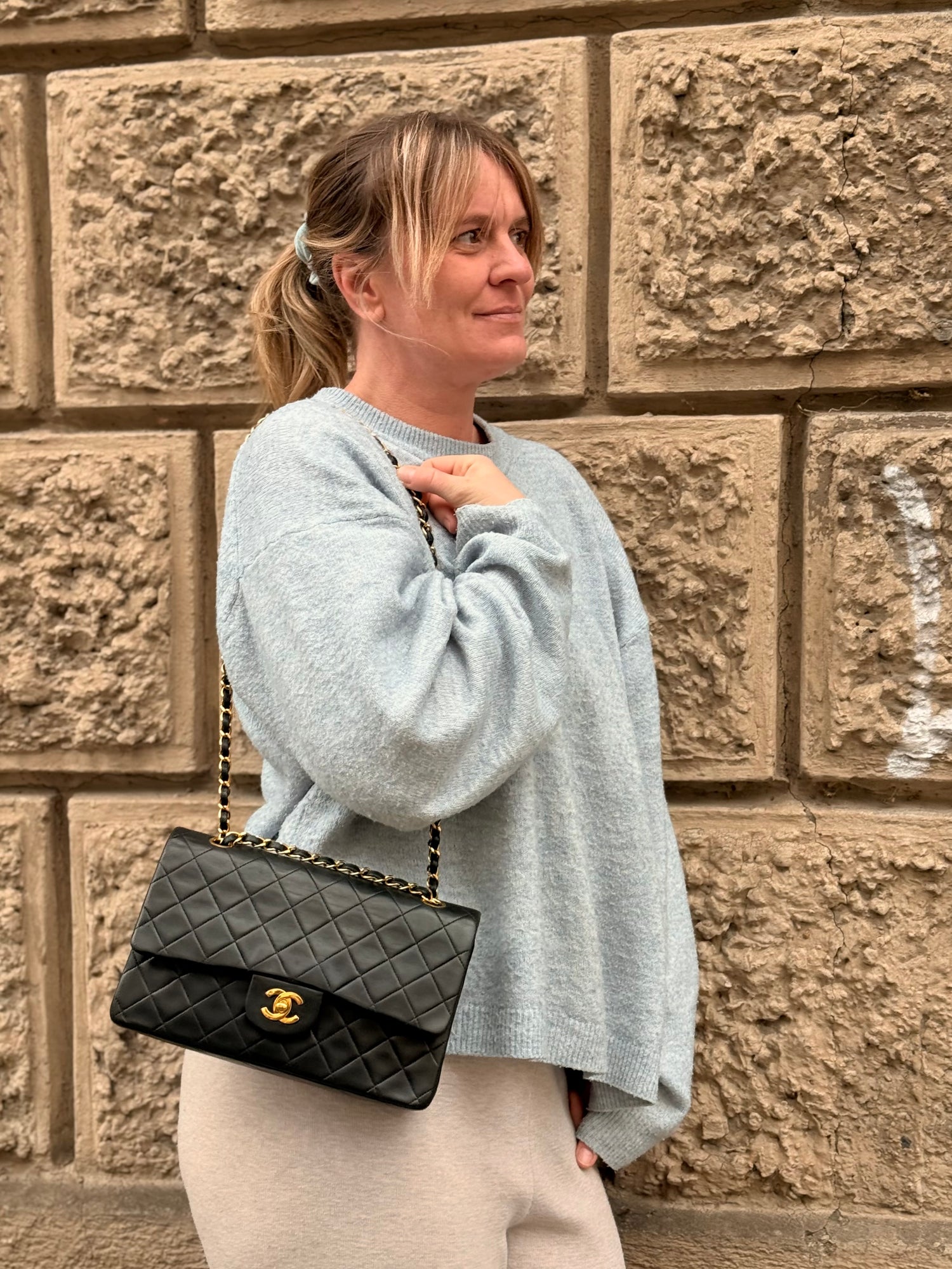 Frau in hellem Pullover und beiger Hose trägt schwarze gesteppte Chanel Handtasche mit goldener Kette vor Sandsteinwand