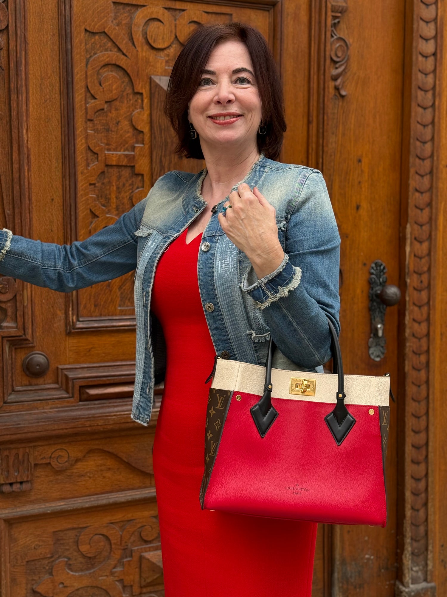 Frau im roten Kleid und Jeansjacke trägt rote Louis Vuitton Tasche mit hellen Details vor geschnitzter Holztür
