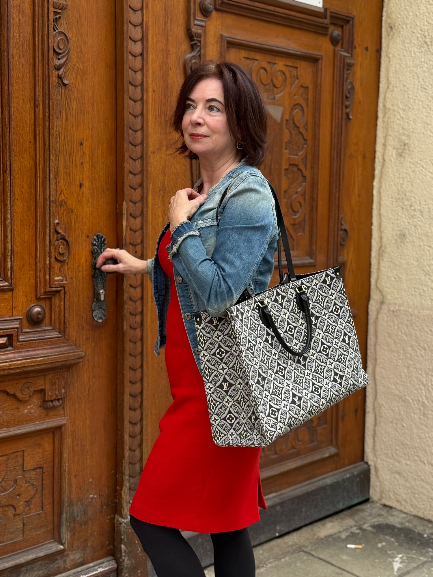 Frau mit rotem Kleid und Jeansjacke trägt große schwarz-weiße Tasche mit Muster vor Holztür