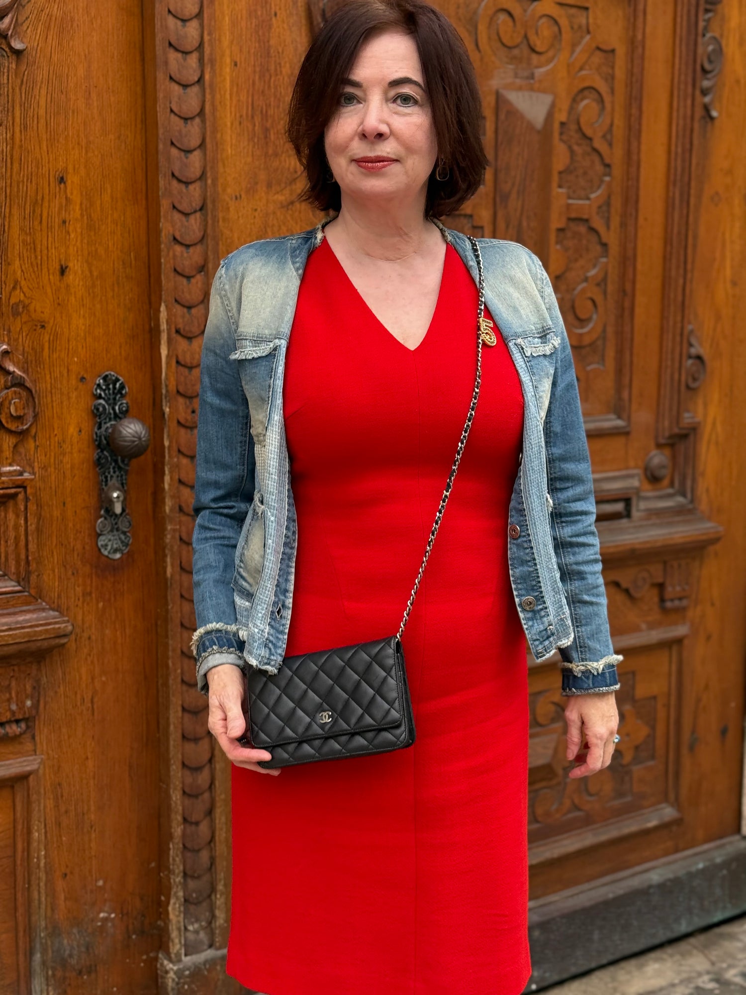Frau in rotem Kleid und Jeansjacke hält schwarze gesteppte CHANEL Handtasche mit goldener Kette vor Holzportal