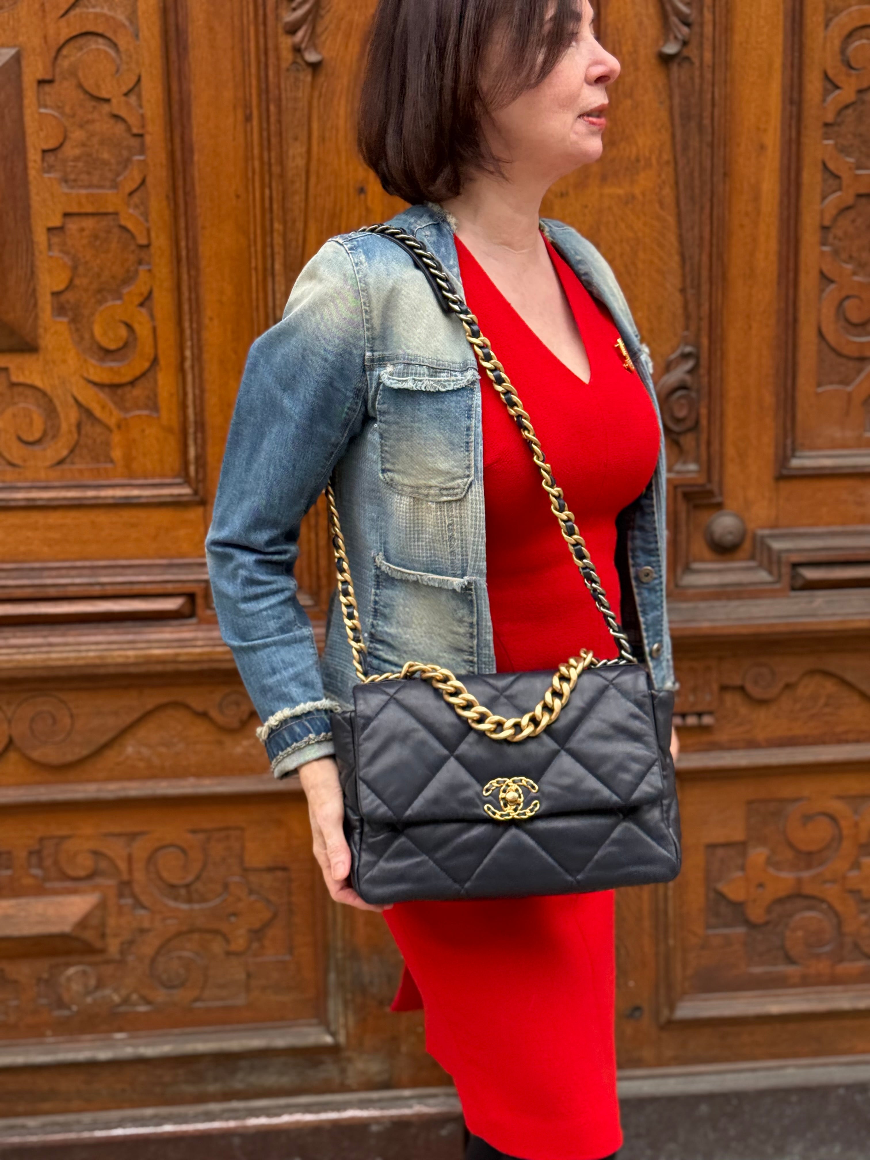 Frau in rotem Kleid und leichter Jeansjacke trägt große schwarze Chanel Handtasche mit goldener Kette vor Holztür