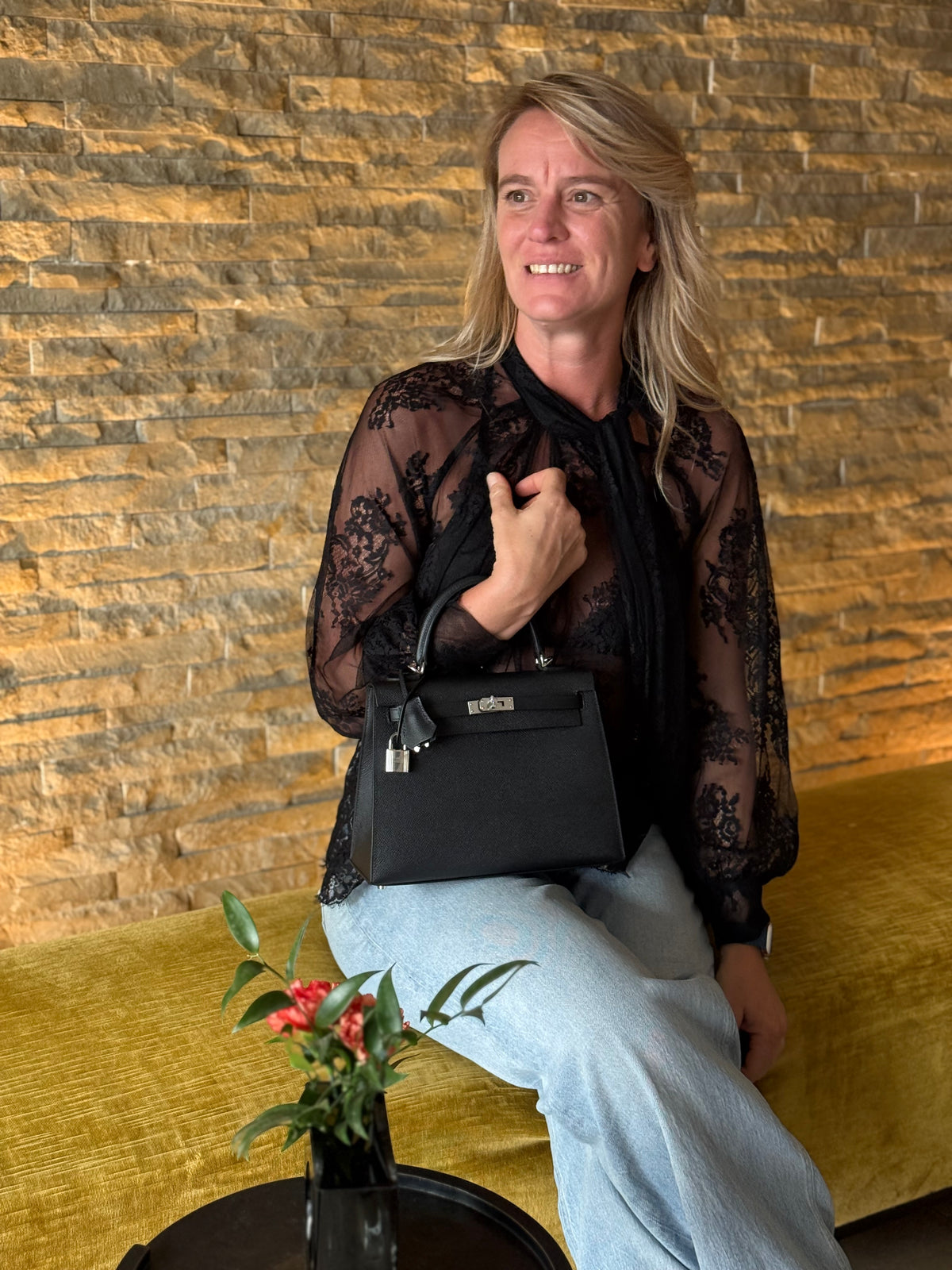 Frau sitzt mit schwarzer Handtasche auf einem Sofa vor einer Natursteinwand und einem Blumenstrauß