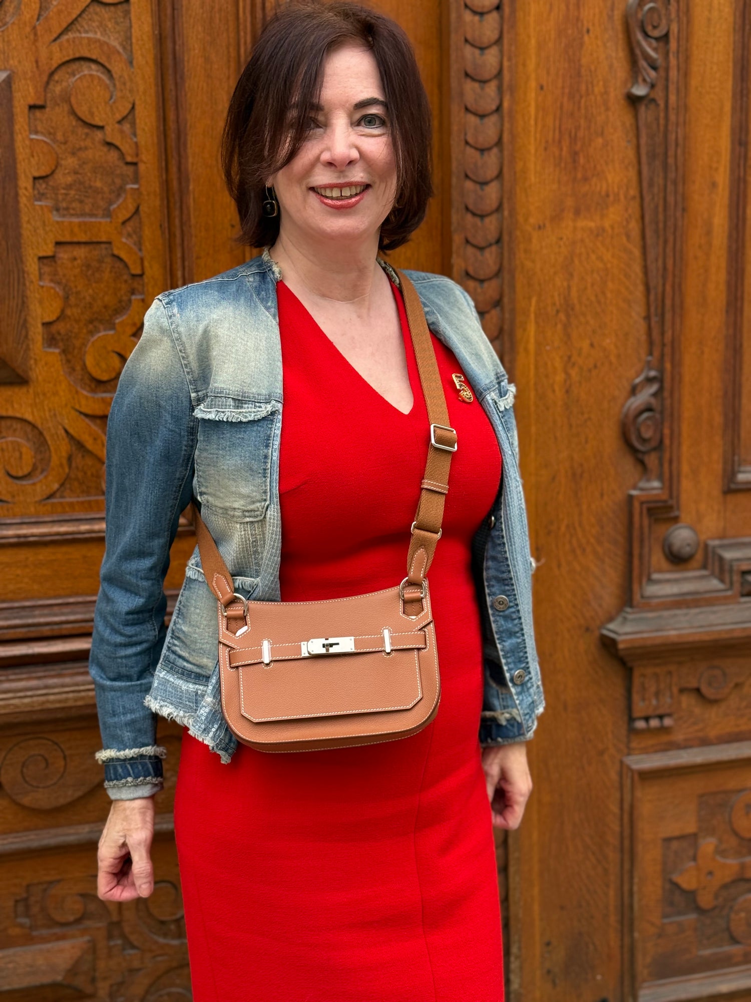 Frau im roten Kleid mit blauer Jeansjacke trägt beige Umhängetasche vor kunstvoll geschnitzter Holztür