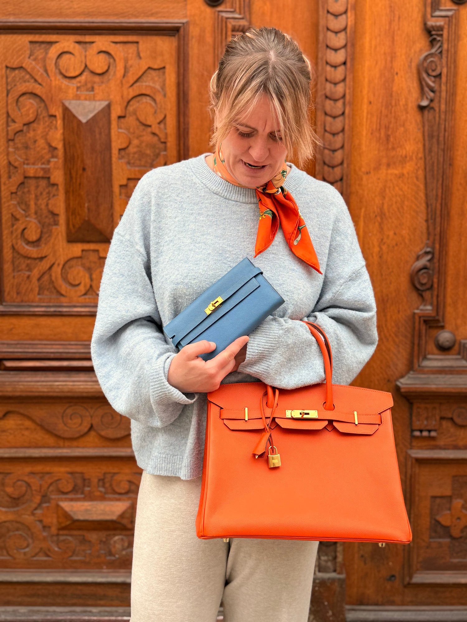 Frau mit grauem Pullover hält orangefarbene Hermès Tasche und blaue Clutch vor geschnitztem Holztor