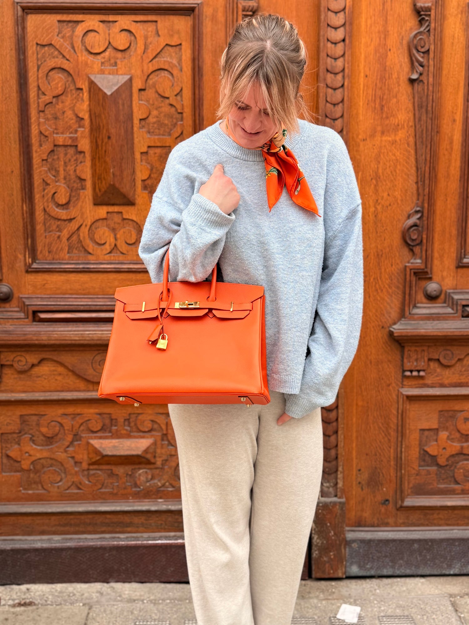 Frau in hellem Pullover mit orangefarbenem Halstuch trägt große orangefarbene Ledertasche vor Holzportal
