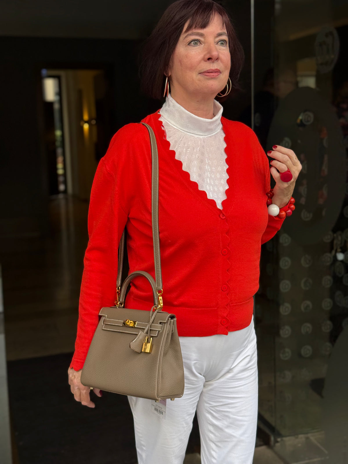 Frau mit roter Strickjacke, weißer Hose und beiger Handtasche vor einer Tür