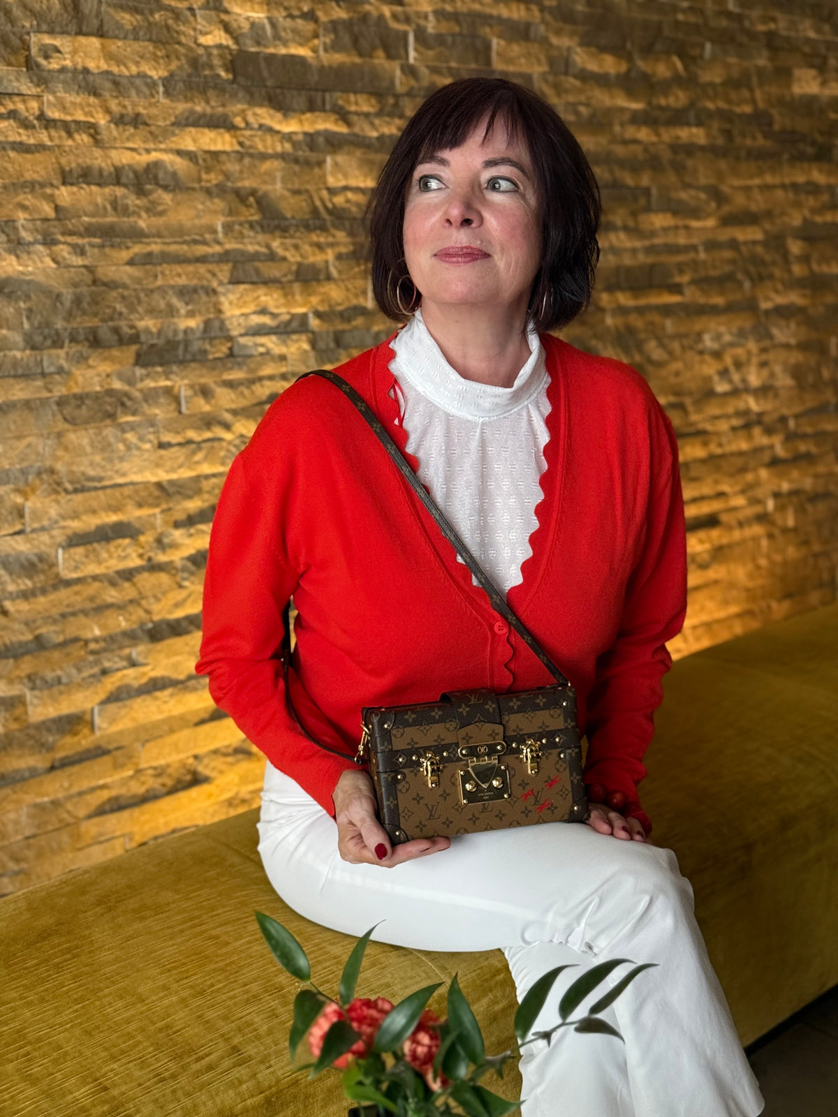 Frau in roter Strickjacke mit brauner Designerhandtasche vor einer rustikalen Steinwand unerquicklich
