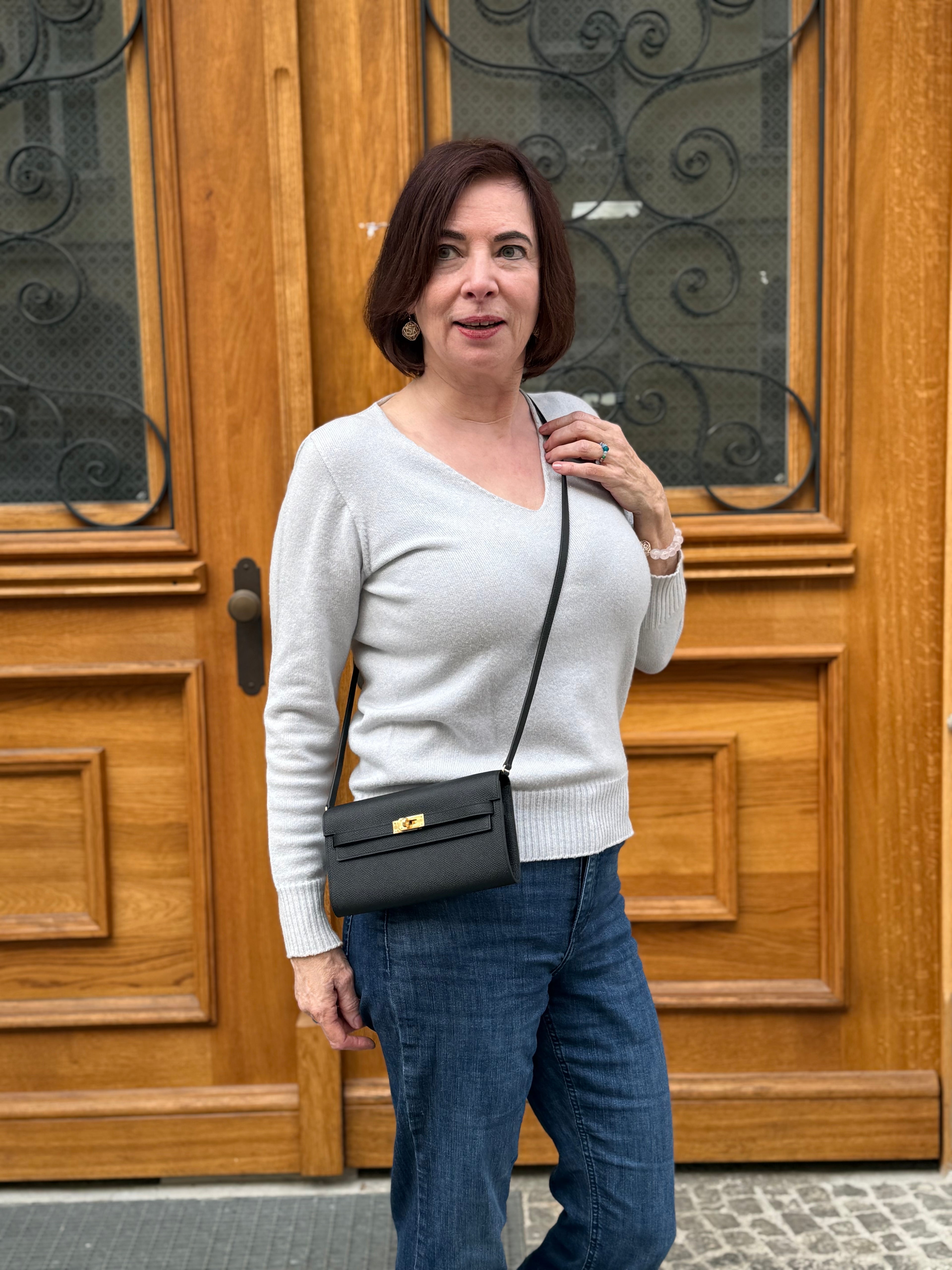 Frau mit braunen Haaren trägt hellen Pullover, blaue Jeans und eine schwarze, rechteckige Umhängetasche vor Holztür