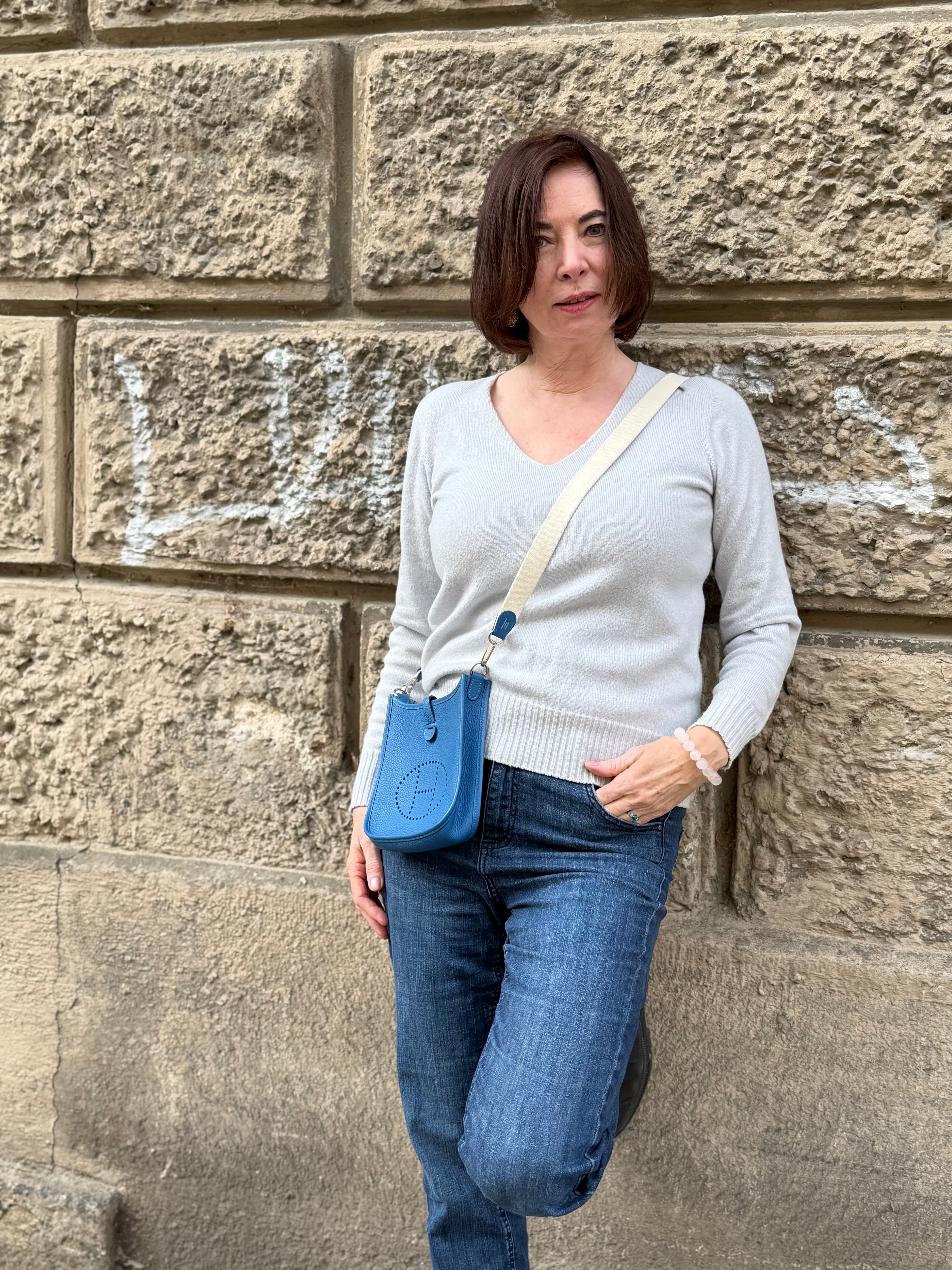 Frau in hellem Pullover und Jeans trägt kleine blaue Ledertasche mit breitem Riemen vor Steinwand mit Graffiti
