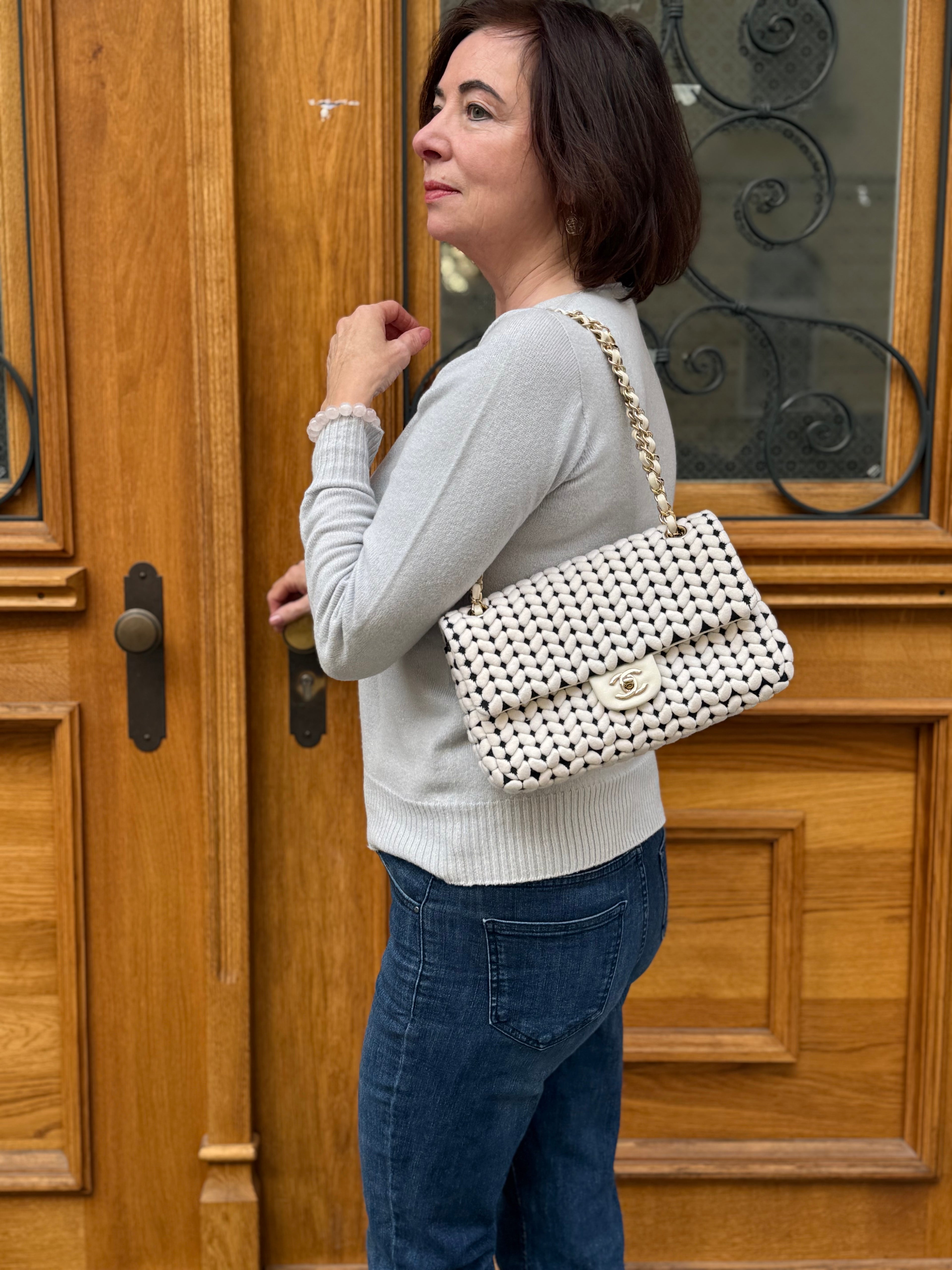 Frau mit hellgrauem Pullover und dunkler Jeans trägt weiße Handtasche mit Goldkette vor Holztür